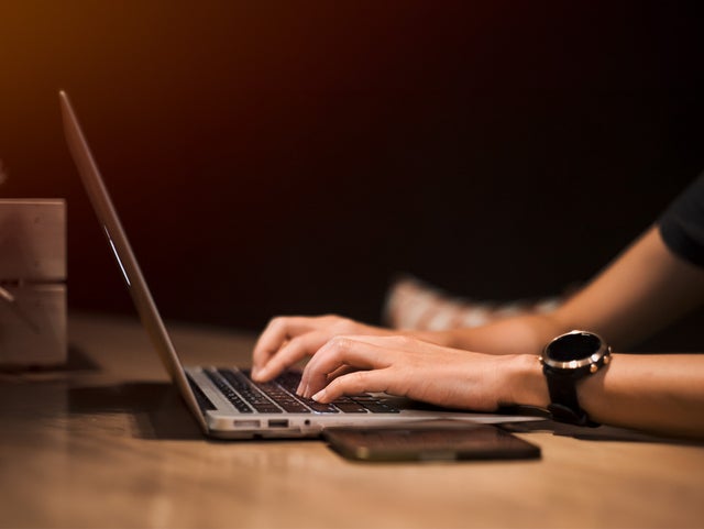 A person types on a laptop in a dimly lit environment, with a smartwatch on their wrist and a smartphone placed on the table beside them. The warm, focused lighting emphasizes productivity and technology use, highlighting a modern workspace or remote work setting. The image conveys themes of digital connectivity, work-life balance, and the integration of smart devices in daily tasks.