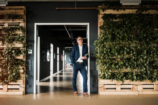 The picture shows a man with short grey hair and glasses in focus. He is wearing a blue suit with a white shirt and brown shoes. While his right hand is in his trouser pocket, he is holding a closed laptop in his other hand. He is leaning against a door frame that leads to a corridor in the background. Several doors can be seen on the left-hand side of the corridor, indicating various offices. To the right and left of the man on the walls are several vertically stacked pallets with green plants running down the pallets, giving the building a modern atmosphere.