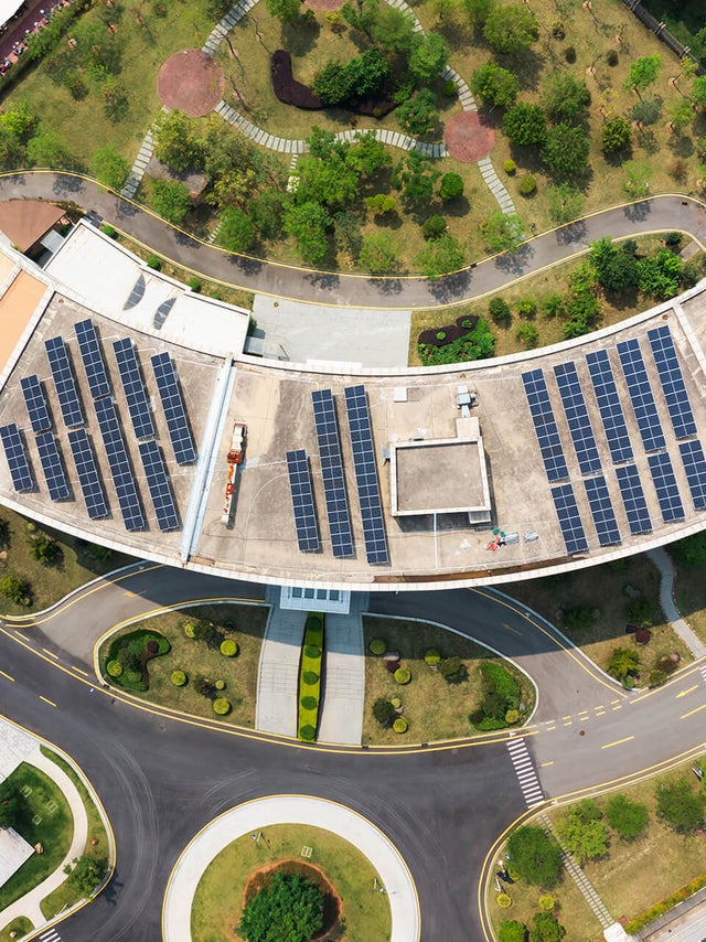 The image shows an aerial view of a modern building complex surrounded by streets, car parks and green spaces. In the centre of the image is a large, curved building with a roof surface that is completely covered with solar panels. The solar panels are arranged in several parallel rows and utilise the entire roof surface. Below the curved building is a wide road that winds around a circular green area with a tree in the centre. This road separates the main building from another large, rectangular building, which is located in the lower left-hand section of the picture. This building has a flat roof and is surrounded by grey paved areas and other green spaces. At the top of the picture is an extensive park with various footpaths running through it. There are numerous trees and shrubs in the park, creating a natural and green environment. At the top right of the picture is a long, straight road that separates the park area from the rest of the grounds. Another road is visible on the left-hand side of the picture, which also has a cycle path and footpath to the left and right of the road. Yellow markings show an entrance to the building complex which is blocked by a barrier.