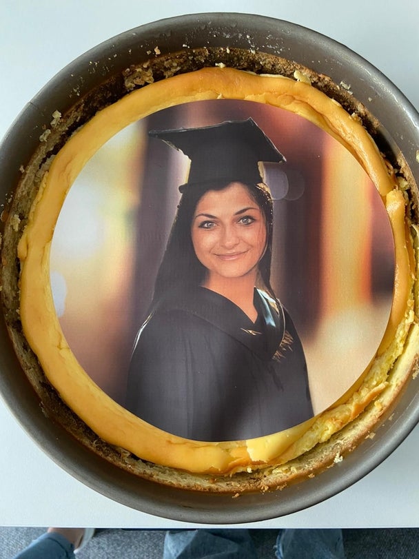 Ein Käsekuchen mit einem essbaren Foto einer jungen Frau in einem schwarzen Abschlussgewand und einem Doktorhut. Das Bild ist mittig auf den Kuchen gedruckt und hebt sich farblich vom Rand ab.