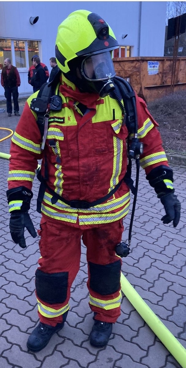 Ein Feuerwehrmann in roter Schutzkleidung mit gelben Reflexstreifen und einem gelben Helm steht auf einem gepflasterten Boden. Er trägt eine Atemschutzmaske und ist mit einem Atemschutzgerät ausgestattet.