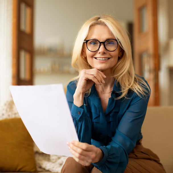 L'image montre une femme assise sur un canapé et lisant la facture de chauffage ista. Elle a de longs cheveux blonds et porte des lunettes noires. Son haut est une chemise bleue et elle porte un pantalon marron. Alors qu'elle tient la facture de chauffage dans sa main gauche, sa main droite est posée sur son menton. Elle regarde directement la caméra. Le canapé beige sur lequel elle est assise s'étend sur tout le tiers inférieur de l'image et comporte une couverture blanche drapée sur le dossier à gauche, ainsi qu'un coussin jaune adossé à celui-ci. À l'arrière-plan, divers meubles évoquent un salon : À droite, on aperçoit un lampadaire et une étagère, et au centre, une porte ouverte en deux parties donnant accès à une autre pièce avec des étagères. L'ensemble de l'arrière-plan est flou.