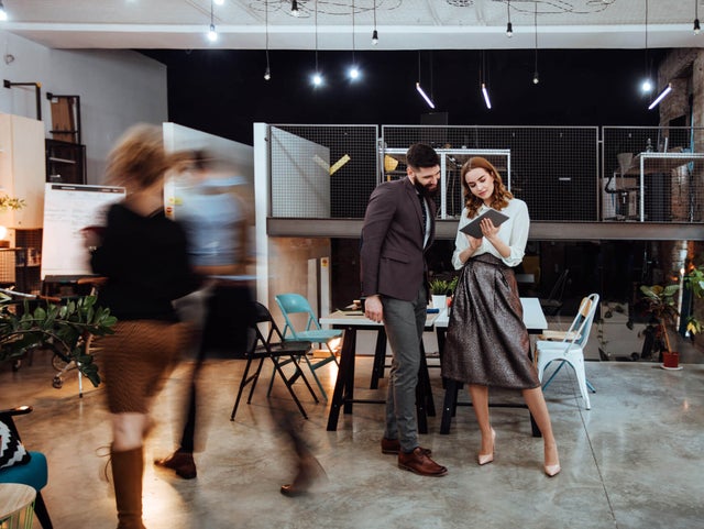 The picture shows a modern, open-plan office space where two people are standing in the center of the room. In the background on the left are cupboards containing files and a presentation board. To the right, you can see two white walls raised close together that do not reach the ceiling of the room. A railing is attached to the right-hand wall, which extends to the right-hand side of the picture and to the wall there. The wall of the railing itself is made of a grey grid and the floor is attached to a steel beam that can be seen from the side. The lattice provides a clear view of the black wall behind it. Two large desks can be seen in the center of this elevation, with two chairs on either side. On the right-hand side, against the brick wall, there is another desk with a chair and a passageway in the wall. In the foreground you can see two people walking towards the cupboards on the left. These two people are only vaguely recognizable as they are in motion and therefore blurry. In front of the railing in the center of the picture are two large tables that have been placed opposite each other and form a large group of tables. There are two chairs to the left and right of the group of tables. Three plants can be seen on the right-hand edge of the picture, their branches protruding into the picture. In the center of the picture, a younger man is standing next to a younger woman, right before the group of tables. The man has short black hair and a black beard and is wearing a black jacket with grey trousers and brown shoes. The woman has open red wavy hair and is wearing a white blouse, a grey metallic skirt and white pumps. They are both smiling while the woman has a tablet in her hand and is presenting the smart metering options from ista to the man.
