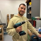 Ein junger Mann in beigem Hoodie hält eine Bohrmaschine in der Hand und steht lächelnd in einer Werkstatt.