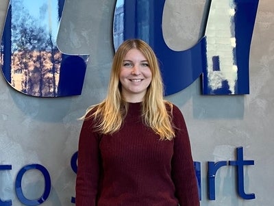 Eine junge Frau steht vor einer Wand mit einem großen blauen Logo, auf dem „ista“ und der Slogan „Switch to Smart“ steht. Sie lächelt und trägt einen dunkelroten Pullover; der Hintergrund ist in neutralen Farben gehalten.