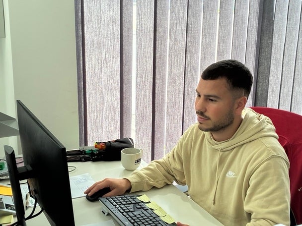 Ein Mann in beigem Hoodie arbeitet konzentriert am Computer in einem hellen Büro mit grauen Vorhängen.