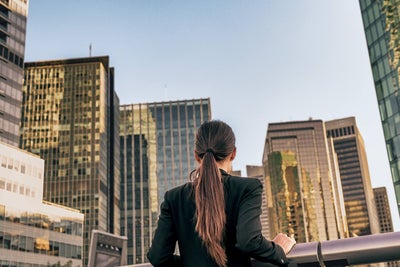 The picture shows a woman standing at a silver railing and looking at a view of many skyscrapers. She has long brown hair tied back in a ponytail and is wearing a black jacket. She is standing with her back to the viewer. The skyscrapers are close together and all have either complete window fronts or many rows of windows stretching through the entire building. The sun shines on the windows of some of the buildings, while the sun shines only partially on the other buildings, because some of the other skyscrapers are casting shadows on them.