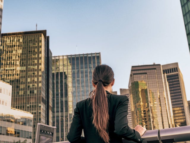 The picture shows a woman standing at a silver railing and looking at a view of many skyscrapers. She has long brown hair tied back in a ponytail and is wearing a black jacket. She is standing with her back to the viewer. The skyscrapers are close together and all have either complete window fronts or many rows of windows stretching through the entire building. The sun shines on the windows of some of the buildings, while the sun shines only partially on the other buildings, because some of the other skyscrapers are casting shadows on them.