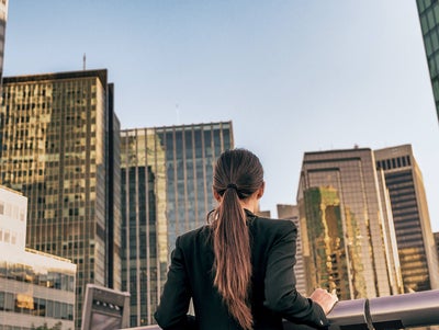 The picture shows a woman standing at a silver railing and looking at a view of many skyscrapers. She has long brown hair tied back in a ponytail and is wearing a black jacket. She is standing with her back to the viewer. The skyscrapers are close together and all have either complete window fronts or many rows of windows stretching through the entire building. The sun shines on the windows of some of the buildings, while the sun shines only partially on the other buildings, because some of the other skyscrapers are casting shadows on them.
