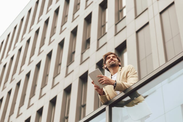 The picture shows a young man leaning against a glass railing and holding a small tablet with a white cover in his hand. He is smiling and looking into the distance. The man has short brown hair and a short brown beard and is wearing a beige suit with a white t-shirt underneath. Since the photo was taken from a frog's-eye view, you can see a tall, modern office building made of light-coloured stone with four rows of large windows in the background.
