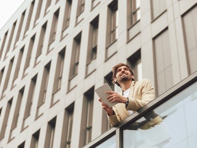 The picture shows a young man leaning against a glass railing and holding a small tablet with a white cover in his hand. He is smiling and looking into the distance. The man has short brown hair and a short brown beard and is wearing a beige suit with a white t-shirt underneath. Since the photo was taken from a frog's-eye view, you can see a tall, modern office building made of light-coloured stone with four rows of large windows in the background.
