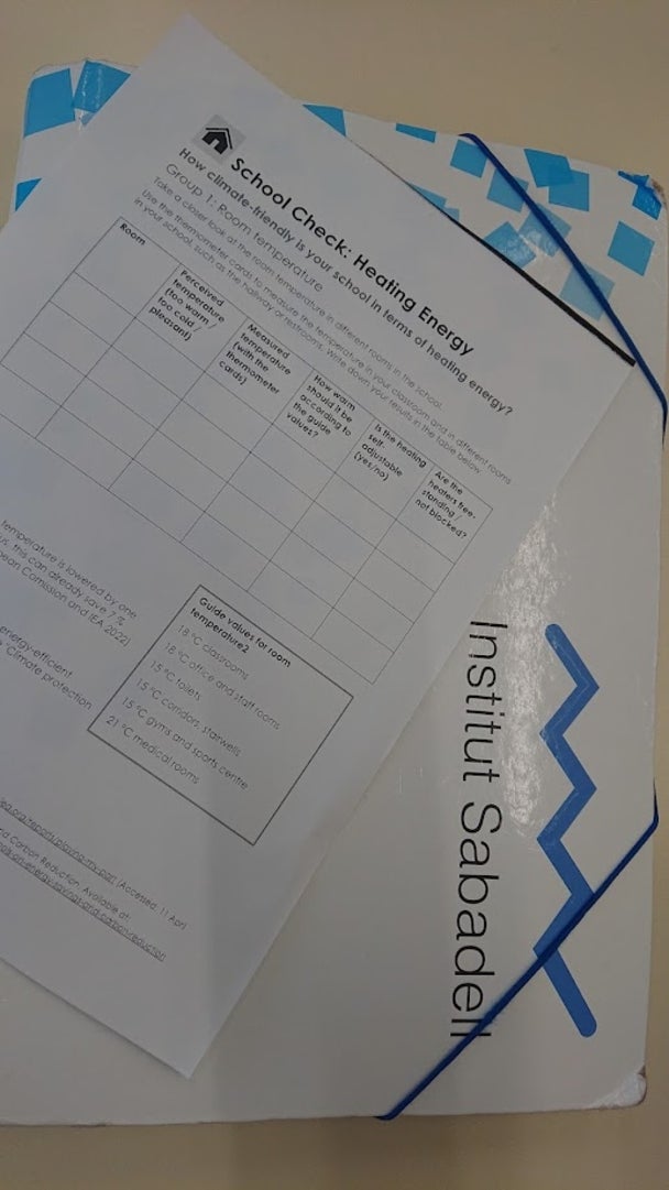 Ein Fragebogen zum Thema „School Check: Heating Energy“ liegt auf einer Mappe mit dem Logo des Instituts Sabadell; der Fragebogen dient zur Bewertung der Raumtemperatur und Energieeffizienz in Schulräumen.