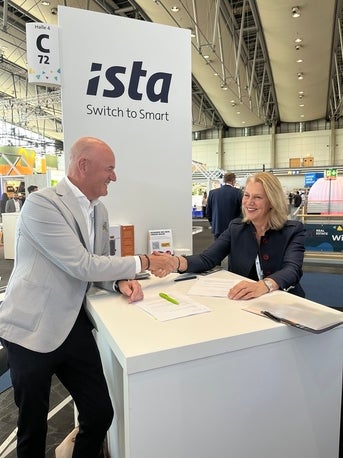Zwei Geschäftsleute, ein Mann und eine Frau, sitzen an einem weißen Tisch auf einer Messe und schütteln sich lächelnd die Hand. Hinter ihnen steht ein großes Schild mit dem Logo der Firma „ista“ und dem Slogan „Switch to Smart“. Auf dem Tisch liegen Vertragsunterlagen und ein grüner Stift. Im Hintergrund sind weitere Messestände und Besucher zu sehen.