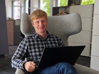 Mann mit blonden Haaren in einem modernen Büro, lächelnd in einem grauen Sessel sitzend, mit einem Laptop auf dem Schoß. Im Hintergrund ein buntes Wandbild mit Naturmotiven.