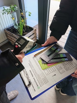 Zwei Personen führen eine Temperaturmessung mit einem Infrarot-Thermometer in einem Schulgebäude durch; eine hält ein Klemmbrett mit Informationsmaterial zum Thema Heizenergie und Klimaschutz.