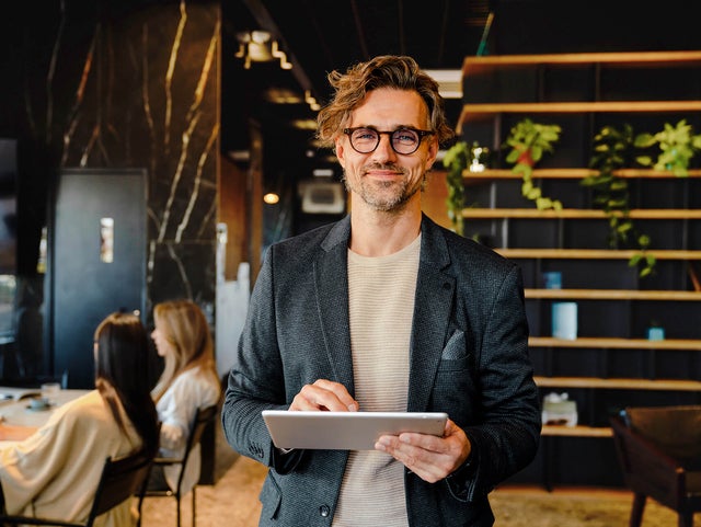 The picture shows a smiling man holding a tablet on which ista's Energy Management Minute View is displayed. The man has brown hair and is wearing black glasses. He is wearing a beige and grey striped shirt and a blue blazer over it. He is standing in the centre of the picture and looking directly into the camera. A modern office can be seen in the background. On the left-hand side of the picture is a wooden shelf in front of a black wall, decorated with long hanging plants and various objects that give the impression of awards. On the right of the picture, two women are sitting next to each other at a white work table covered with various work items. The woman in front, whose long black hair and beige loose top are visible, rests her arms on the table and has turned her head towards her colleague, so that only the back of her head is visible. The woman next to her has long blonde hair and is wearing a light blue shirt. Her side profile is recognisable. The entire background is blurred so that the focus is on the smiling man with the tablet.