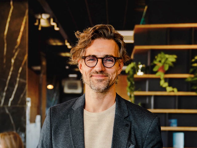 The picture shows a smiling man holding a tablet on which ista's Energy Management Minute View is displayed. The man has brown hair and is wearing black glasses. He is wearing a beige and grey striped shirt and a blue blazer over it. He is standing in the centre of the picture and looking directly into the camera. A modern office can be seen in the background. On the left-hand side of the picture is a wooden shelf in front of a black wall, decorated with long hanging plants and various objects that give the impression of awards. On the right of the picture, two women are sitting next to each other at a white work table covered with various work items. The woman in front, whose long black hair and beige loose top are visible, rests her arms on the table and has turned her head towards her colleague, so that only the back of her head is visible. The woman next to her has long blonde hair and is wearing a light blue shirt. Her side profile is recognisable. The entire background is blurred so that the focus is on the smiling man with the tablet.
