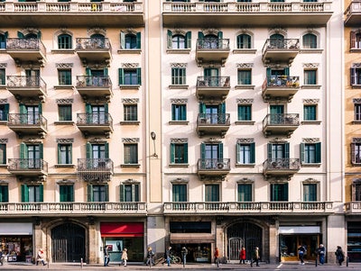 The image depicts a bustling urban street scene featuring a classic European-style apartment building. The building's facade is characterized by elegant architectural details, including wrought iron balconies, green shutters, and ornate window frames. The structure stands four stories tall, with retail shops on the ground floor, marked by large display windows and signage. Pedestrians are seen walking along the sidewalk, some engaged in conversation, while others appear to be in transit. The overall atmosphere is vibrant, reflecting a lively city environment.