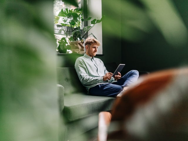 The picture shows a young man sitting comfortably on a couch, busy with a tablet. In the foreground, a plant and a chair are intentionally blurred to focus on the man's interaction with the tablet. This scene creates a cozy and relaxed atmosphere and emphasizes the use of modern technology in a casual home environment.