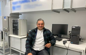 Ein Mann sitzt auf einem Hocker in einem modernen Prüfraum mit einem Test-Tunnel im Hintergrund. Er trägt ein kariertes Hemd über einem weißen T-Shirt und blickt freundlich in die Kamera.