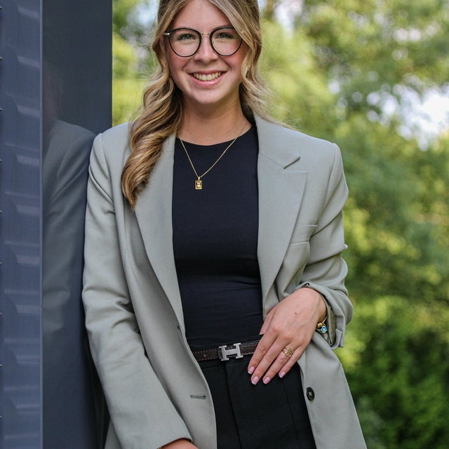 Eine junge Frau mit blonden, gewellten Haaren und Brille steht lächelnd vor einem modernen Gebäude im Freien. Sie trägt einen hellgrauen Blazer, ein schwarzes Oberteil und eine schwarze Hose mit Gürtel. Im Hintergrund sind Bäume und ein Garten sichtbar.