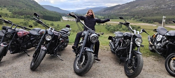 Inga Seimet sitzt begeistert auf einem schwarzen Motorrad mit hohen Lenkern, umgeben von mehreren Motorrädern. Sie trägt eine schwarze Motorradjacke und hat die Arme weit ausgebreitet. Der Hintergrund zeigt eine malerische Landschaft mit grünen Hügeln, Bäumen und einem Fluss im Tal.