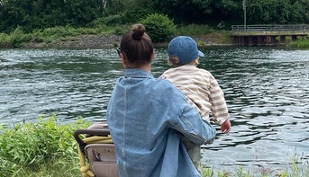 Eine Frau, die einen Dutt trägt und eine Sonnenbrille aufhat, steht mit ihrem kleinen Kind am Flussufer. Beide blicken auf das Wasser. Die Frau trägt eine Jeansjacke, das Kind eine gestreifte Jacke und eine blaue Mütze. Neben ihnen steht ein Kinderwagen.