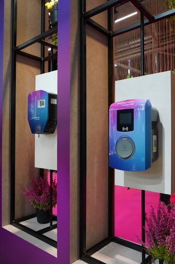 Zwei moderne Wallboxen für Elektrofahrzeuge in einem stilvollen Rahmen mit violetten und blauen Farbtönen, dekoriert mit Pflanzen und pinkem Hintergrund.