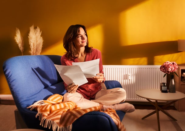 Eine brünette Frau sitzt im Schneidersitze auf ihrer blauen Couch im Wohnzimmer. In den Händen hält Sie zwei Seiten Papier, Seiten aus der Heizkostenabrechnung von ista. Sie blickt zufrieden Richtung Fenster. 