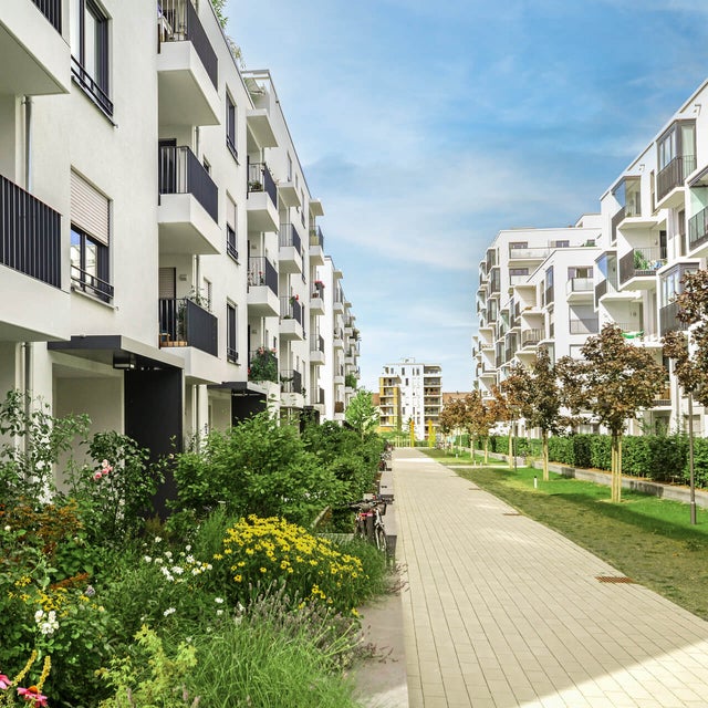 The picture shows a modern residential complex with several white, multi-storey residential buildings standing on the left and right side of a paved path. The buildings have balconies with dark railings, and some balconies are decorated with plants and furniture. A wide, paved footpath stretches between the buildings and continues into the distance. On the left side of the path are lush green areas and colourful flowerbeds that embellish the space between the buildings. On the right-hand side of the path, young trees are planted at regular intervals, forming a green avenue together with manicured lawns. Other buildings can be seen in the background, completing the urban surroundings of the residential complex. The sky is clear and blue, indicating a sunny day. Overall, the picture conveys a calm and pleasant living atmosphere in a modern, well-maintained environment.
