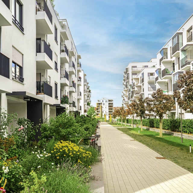 The picture shows a modern residential complex with several white, multi-storey residential buildings standing on the left and right side of a paved path. The buildings have balconies with dark railings, and some balconies are decorated with plants and furniture. A wide, paved footpath stretches between the buildings and continues into the distance. On the left side of the path are lush green areas and colourful flowerbeds that embellish the space between the buildings. On the right-hand side of the path, young trees are planted at regular intervals, forming a green avenue together with manicured lawns. Other buildings can be seen in the background, completing the urban surroundings of the residential complex. The sky is clear and blue, indicating a sunny day. Overall, the picture conveys a calm and pleasant living atmosphere in a modern, well-maintained environment.