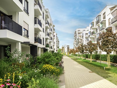 The picture shows a modern residential complex with several white, multi-storey residential buildings standing on the left and right side of a paved path. The buildings have balconies with dark railings, and some balconies are decorated with plants and furniture. A wide, paved footpath stretches between the buildings and continues into the distance. On the left side of the path are lush green areas and colourful flowerbeds that embellish the space between the buildings. On the right-hand side of the path, young trees are planted at regular intervals, forming a green avenue together with manicured lawns. Other buildings can be seen in the background, completing the urban surroundings of the residential complex. The sky is clear and blue, indicating a sunny day. Overall, the picture conveys a calm and pleasant living atmosphere in a modern, well-maintained environment.