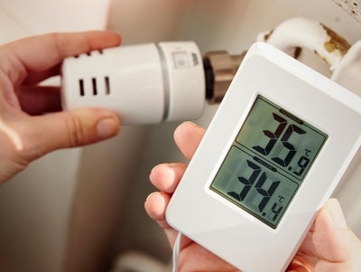 Ein Hand reguliert das Heizugsthermostat, während eine andere ein Messgerät mit Temperatur und Luftfeuchtigkeit hält. 