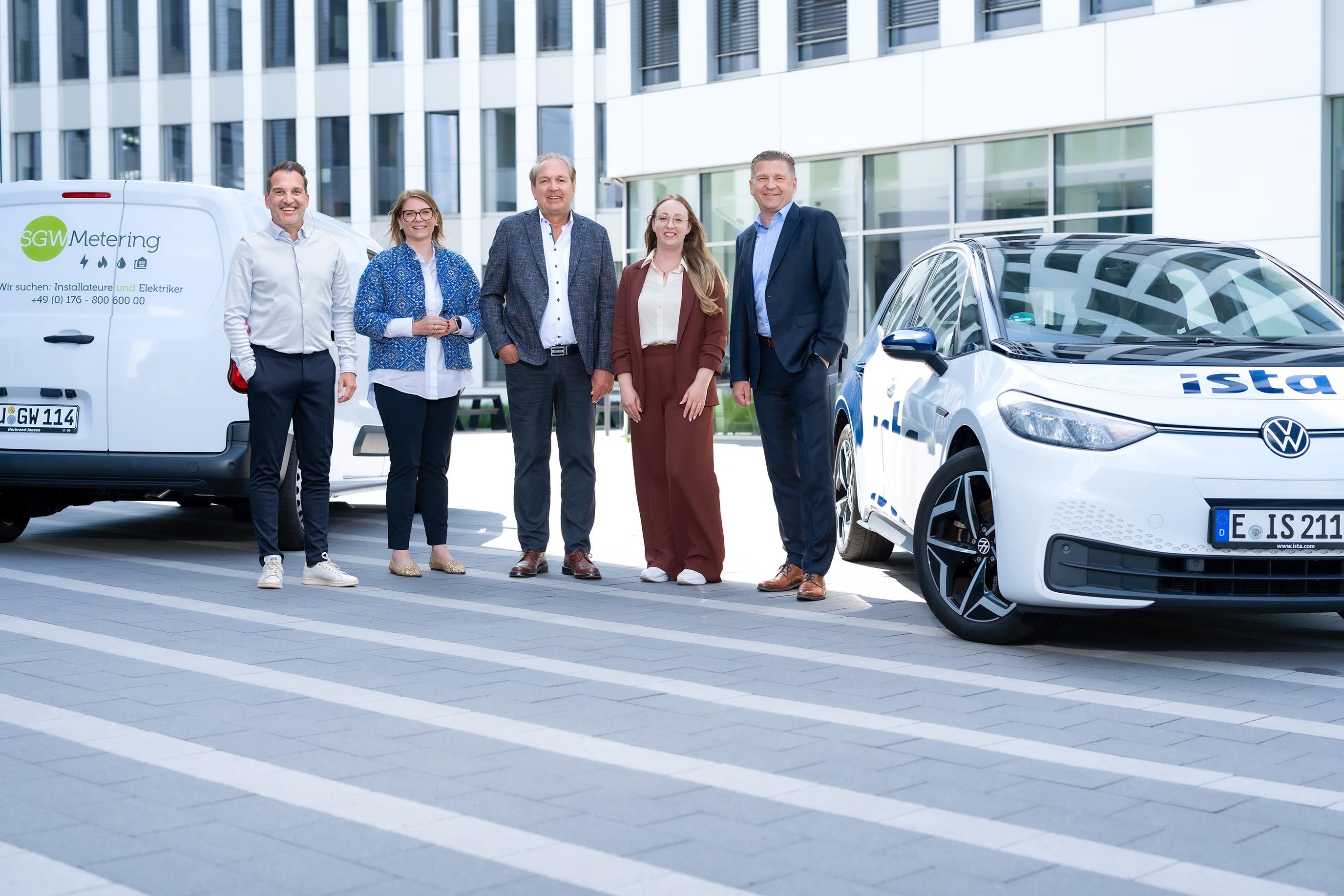 Fünf Personen stehen lächelnd nebeneinander vor einem modernen Bürogebäude. Links ist ein weißer Transporter mit der Aufschrift „SGW Metering“ zu sehen, rechts ein weiß-blaues Elektroauto von „ista“. Die Gruppe wirkt geschäftlich gekleidet und symbolisiert eine Kooperation zwischen den beiden Unternehmen.