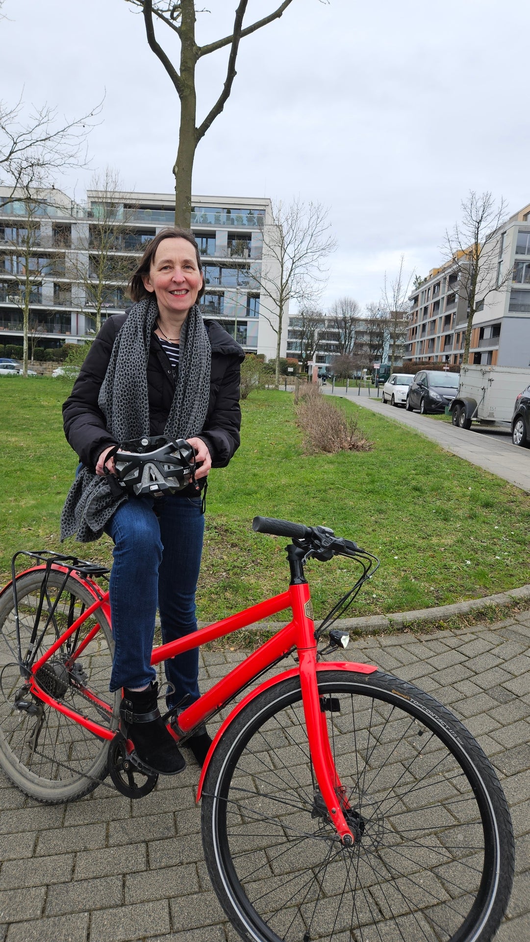 Unsere Kollegin Michaela Hünemeier sitzt auf ihrem roten Fahrrad, trägt ihren Helm in der Hand und lächelt in die Kamera. Hinter ihr sieht man eine grüne Wiese, im weiteren Hintergrund stehen Wohnhäuser.