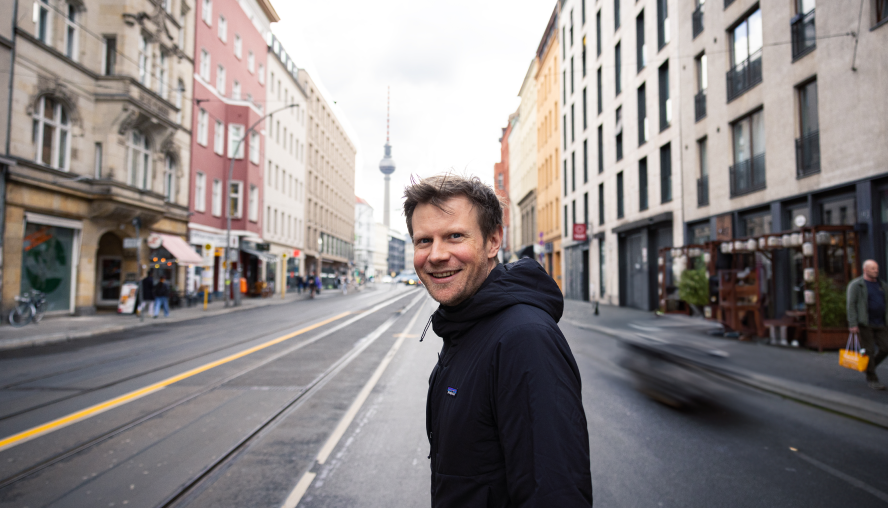 Unser Kollege Bastian Pfahl steht auf einer Straße in Berlin. In der Flucht erkennt man den Berliner Fernsehturm. Er lächelt freundlich in die Kamera. 