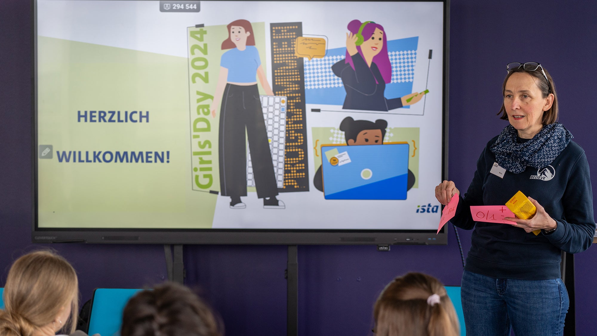 Eine Frau hält einen Vortrag in einem Konferenzraum, steht neben einem großen digitalen Bildschirm, der eine Willkommensbotschaft für den Girls Day 2024 anzeigt. Sie hält und betrachtet rosa und gelbe Haftnotizen, während sie ein nicht im Bild sichtbares Publikum anspricht.