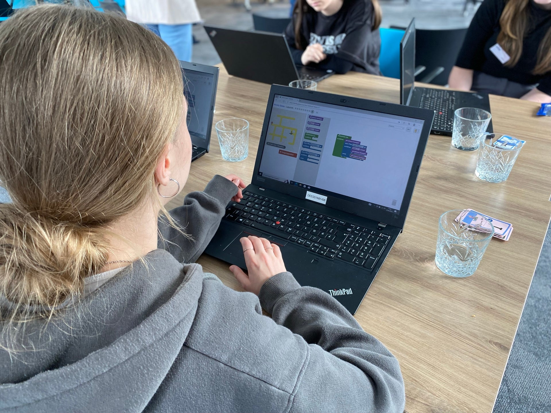 Ein Foto über die Schulter einer Person, die vor einem Laptop sitzt und programmiert. Auf dem Bildschirm sind farbige Codeblöcke zu sehen. Im Hintergrund sitzen weitere Personen, die ebenfalls auf Laptops arbeiten. Auf dem Tisch stehen Wassergläser und ein Stapel Karten.