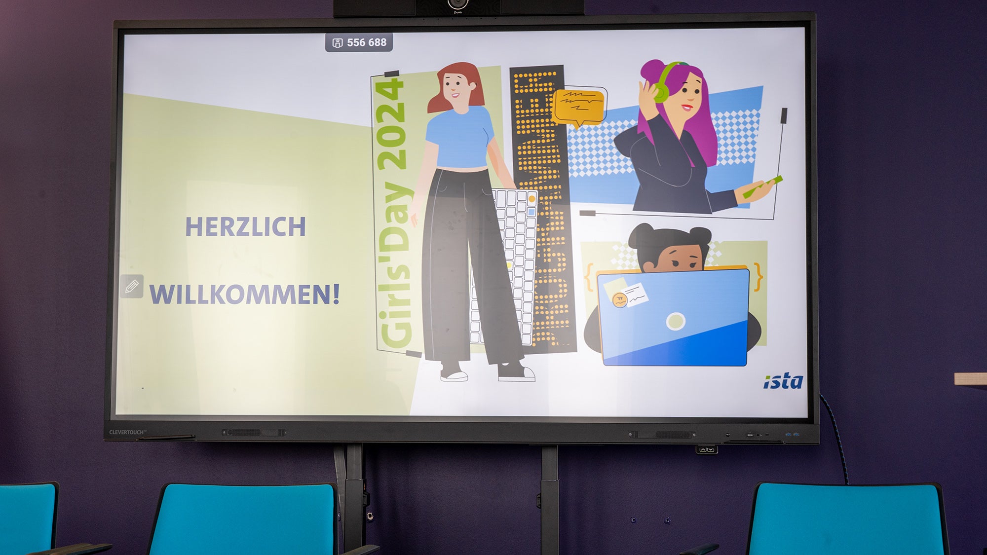 Ein Präsentationsbildschirm in einem Konferenzraum zeigt eine Willkommensfolie mit der Aufschrift "HERZLICH WILLKOMMEN!" für den Girls Day 2024, mit Illustrationen von Mädchen, die sich mit Technologie und Wissenschaft beschäftigen.