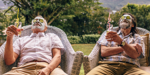 Teaser für Freunde werben Freunde: Zwei Männer sitzen zurückgelehnt auf Rattansesseln im Garten. Sie haben jeweils ein mit Strohhalm in der Hand und haben Gesichtsmasken mit Gurkenscheiben im Gesicht.