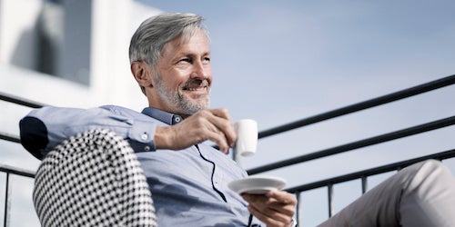 Ein Mann im blauen Hemd sitzt mit einer Tasse Kaffee auf einem Balkon. Er lehnt sich entspannt zurück und sieht zufrieden aus. 