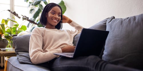 Eine Frau in hellem Pullover sitzt entspannt auf einer Couch. Sie guckt fröhlich und arbeitet an einem Laptop, den Sie auf den Beinen stehen hat. 