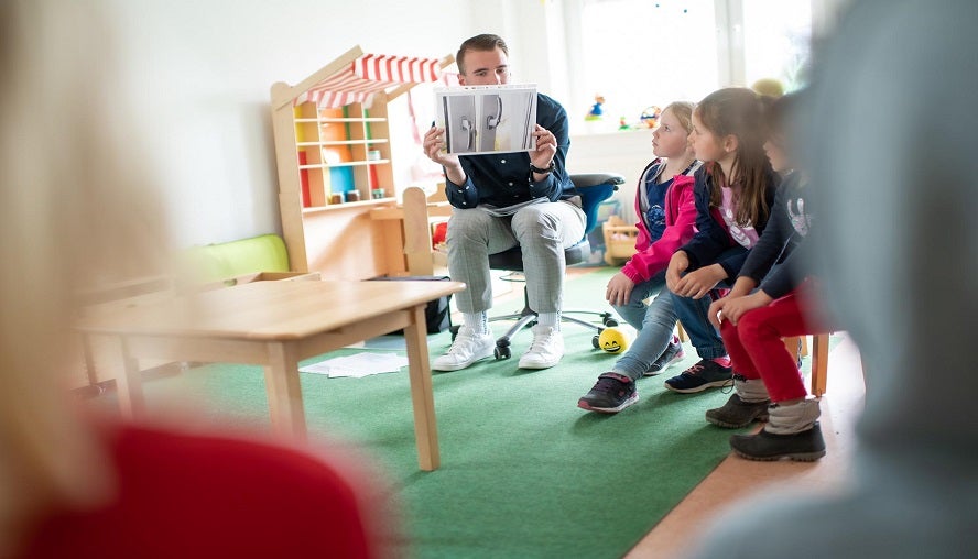 Das Foto ist aus der Perspektive eines Betrachters aufgenommen, da es zwischen zwei Personen entstanden ist. Der Fokus liegt auf einer Gruppe von Personen, die sich in einem Kindergartenraum mit Spielzeugen und einem Holztisch befinden. Davon ein Mann und  drei kleine Mädchen. Der Mann sitzt auf einem Stuhl und hält dabei ein Foto vor seinem Gesicht, sodass die Mädchen das Bild sehen können. Diese sitzen direkt neben ihm und ihre Blicke sind interessiert auf das Bild gerichtet.