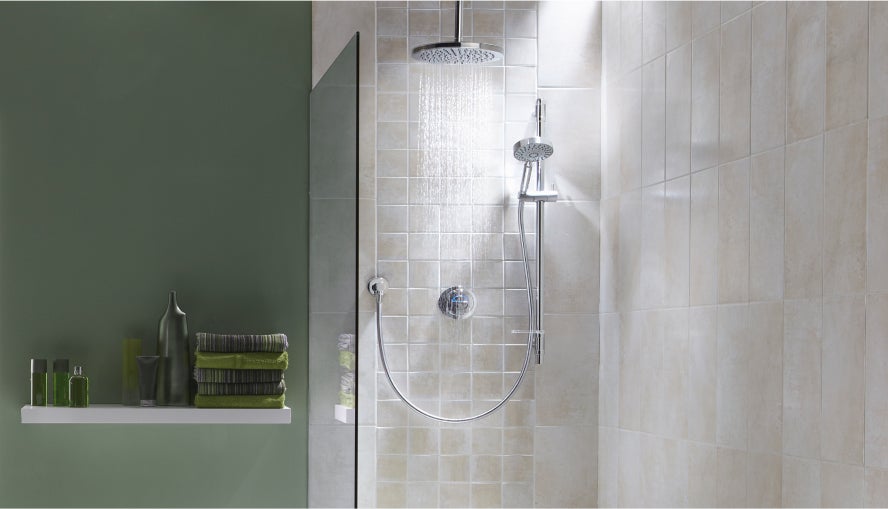 Im Bild sieht man ein Badezimmer. Darin ist eine freiliegende Dusche mit zwei Duschköpfen zu sehen. Das eine ist an der Decke befestigt und das andere an einem Schlauch, der an der Wand angebracht ist. Die Wände da sind aus weißen Fliesen. Die  Dusche wird mit einer Glaswand abgetrennt. Daneben ist eine grüne Wand zu sehen, an der ein weißes Regal befestigt ist, wo grüne Duschartikel und Dekoration zu sehen sind, darunter sowas wie Handtücher oder Tuben.