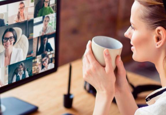 Eine Frau sitzt mit einer Tasse in der Hand an ihrem Schreibtisch. Auf dem Monitor sind mehrere Teilnehmer in einem Videocall zu sehen. 