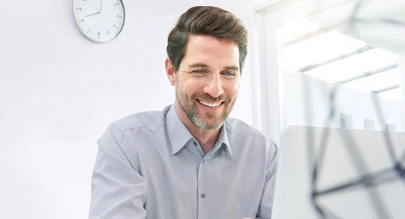 Ein Mann mit dunklen Haaren im grauen Hemd sitzt vor seinem PC. Er scheint in einem Büro zu sein. Im Hintergrund erkennt man eine Uhr.